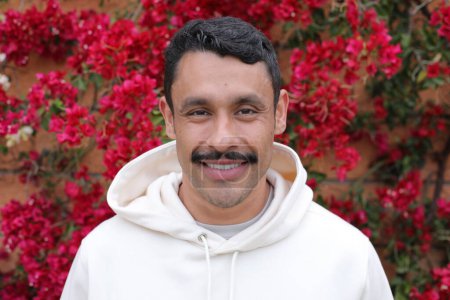 Photo for Portrait of handsome hispanic man with mustache in white hoodie smiling in front of wall of red flowers - Royalty Free Image