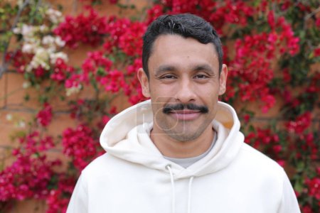Téléchargez les photos : Portrait d'un bel homme hispanique avec moustache à capuche blanche souriant devant un mur de fleurs rouges - en image libre de droit