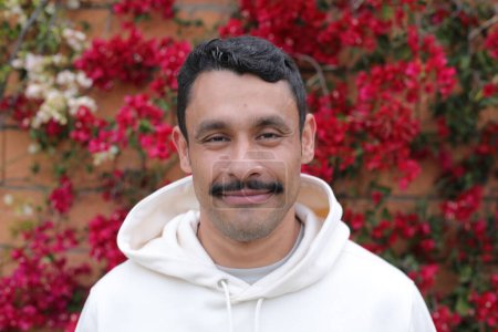 Téléchargez les photos : Portrait d'un bel homme hispanique avec moustache à capuche blanche souriant devant un mur de fleurs rouges - en image libre de droit