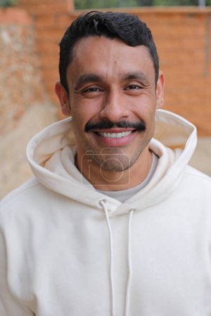Téléchargez les photos : Portrait d'un bel homme hispanique avec moustache en sweat à capuche blanc souriant à la caméra - en image libre de droit