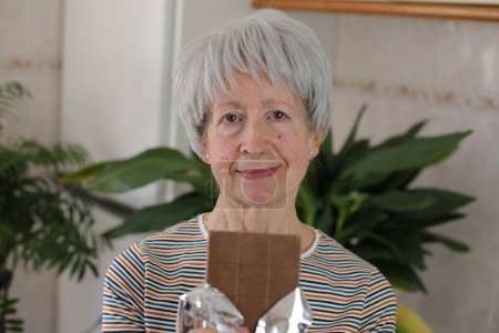 Photo for Portrait of mature grey haired woman holding milk chocolate at home - Royalty Free Image