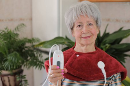 Téléchargez les photos : Portrait de femme mature aux cheveux gris avec couvercle chauffant électrique à la maison - en image libre de droit