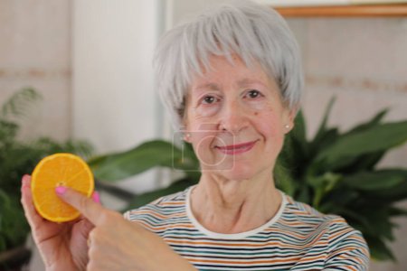 Téléchargez les photos : Portrait d'une femme mûre aux cheveux gris tenant la moitié de l'orange à la maison - en image libre de droit