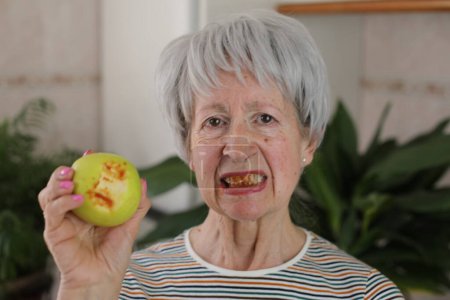 Photo for Portrait of mature grey haired woman with bleeding teeth eating green apple at home - Royalty Free Image