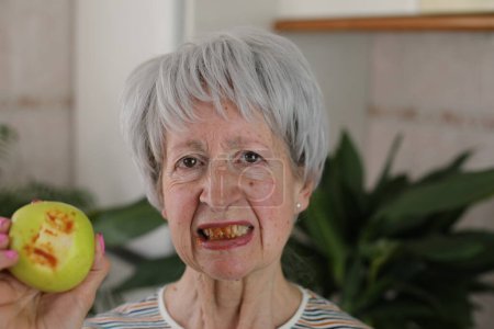 Foto de Retrato de mujer madura de pelo gris con dientes sangrantes comiendo manzana verde en casa - Imagen libre de derechos