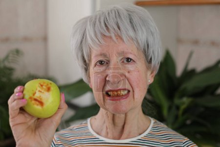 Foto de Retrato de mujer madura de pelo gris con encías sangrantes comiendo manzana verde en casa - Imagen libre de derechos