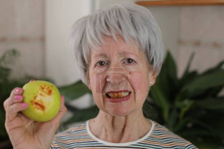 Foto de Retrato de mujer madura de pelo gris con encías sangrantes comiendo manzana verde en casa - Imagen libre de derechos