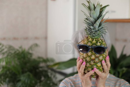 Téléchargez les photos : Portrait d'une femme aux cheveux gris mature tenant un drôle d'ananas avec des lunettes de soleil devant son visage à la maison - en image libre de droit