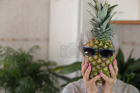Photo for Portrait of mature grey haired woman holding funny pineapple with sunglasses in front of her face at home - Royalty Free Image