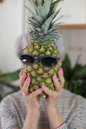 Téléchargez les photos : Portrait d'une femme aux cheveux gris mature tenant un drôle d'ananas avec des lunettes de soleil devant son visage à la maison - en image libre de droit
