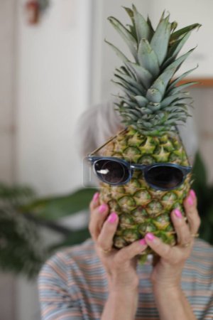 Téléchargez les photos : Portrait d'une femme mûre aux cheveux gris tenant un ananas mûr avec des lunettes de soleil devant son visage à la maison - en image libre de droit