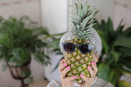 Photo for Portrait of mature grey haired woman holding ripe pineapple with sunglasses in front of her face at home - Royalty Free Image