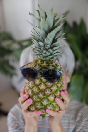 Photo for Portrait of mature grey haired woman holding pineapple with sunglasses in front of her face at home - Royalty Free Image