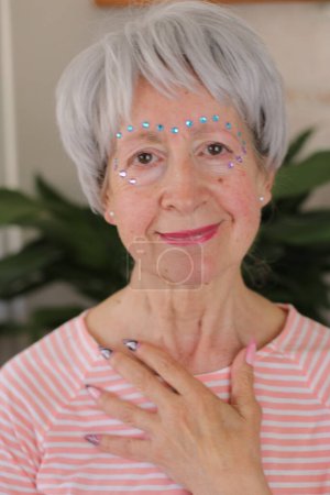 Foto de Retrato de mujer madura de pelo gris con gemas brillantes en la cara y uñas pintadas en casa - Imagen libre de derechos
