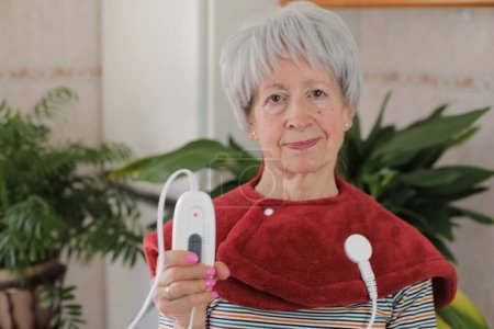 Foto de Mujer mayor con dolor de cuello usando almohadilla eléctrica calentada - Imagen libre de derechos