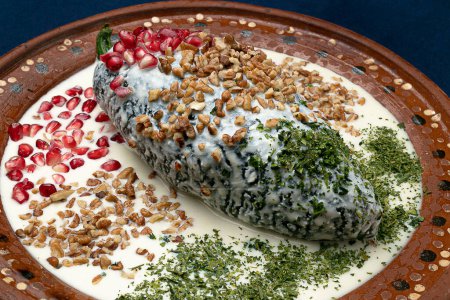 Foto de Comida típica mexicana llamada chiles en nogada adornada con granada, nueces y perejil en un plato tradicional de arcilla sobre una mesa de mantel azul profundo - Imagen libre de derechos
