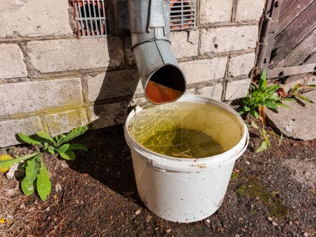 View of plastic bucket outdoors for collecting and storing rainwater from roof run-off in urban city environment with brick wall and asphalt in the frame. Rainwater harvesting