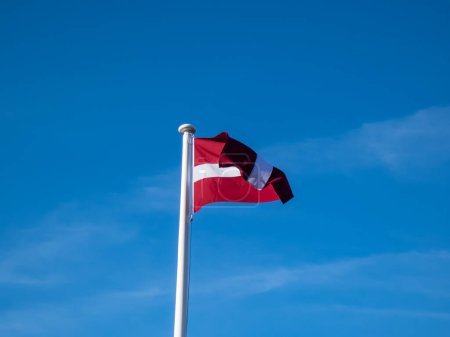 La bandera nacional de Letonia en el viento con el cielo azul nublado. Los colores exactos de la bandera letona - carmín rojo con la raya blanca horizontal. Bandera de Letonia independiente rojo-blanco-rojo