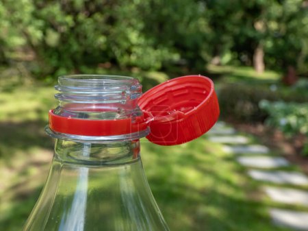 Nahaufnahme des roten Plastikflaschenverschlusses, der an der Flasche befestigt ist, mit extra Plastikstreifen im hellen Sonnenlicht mit grünem Hintergrund