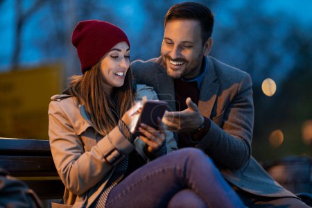 Photo for Happy young couple sitting in a park bench and using smart phone. Enjoying at night. - Royalty Free Image