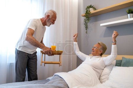 Foto de Desayuno en la cama. Hombre mayor trayendo el desayuno a su esposa en el dormitorio. - Imagen libre de derechos