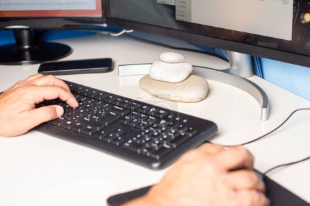 Table de bureau avec des mains travaillant avec un ordinateur et quelques pierres de plage sur la table formant une tour Apacheta.