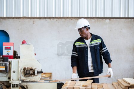 Foto de Hombre americano africano con uniforme de seguridad y sombrero de trabajo duro en máquinas eléctricas de lijado de madera en el taller de fabricación de madera. Hombre carpintero trabajador madera almacén industria. - Imagen libre de derechos