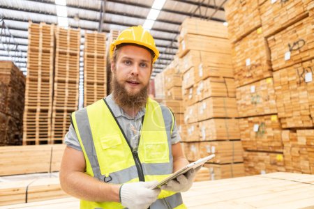 Téléchargez les photos : Travailleur Homme avec une barbe portant un casque de sécurité uniforme jaune à l'aide d'une tablette de travail vérifier la qualité en bois à la fabrication en bois. Homme charpentier ouvrier bois entrepôt industrie. - en image libre de droit
