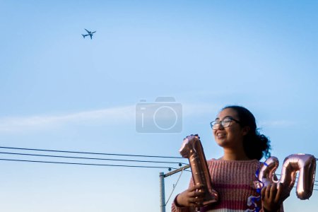 Frau lächelt und hält Geburtstagsballons auf einer Terrasse in der lateinischen Stadt mit einem elektrischen Turm bei Sonnenuntergang und einem Flugzeug im Hintergrund