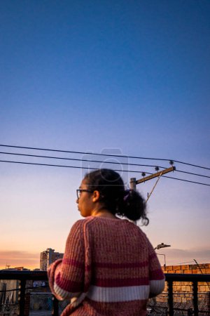 Mujer de pie y con suéter rosa en la terraza de la ciudad latina con una torre eléctrica al atardecer