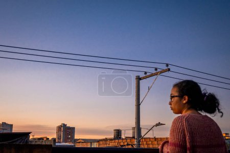 Mujer de pie y con suéter rosa en la terraza de la ciudad latina con una torre eléctrica al atardecer