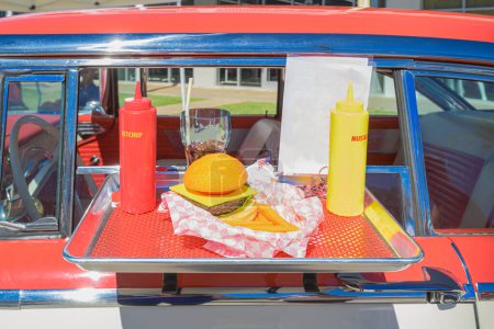"Klassischer Drive-in-Nostalgie Ein rot-weißes Oldtimer-Auto mit einem Futtertablett am Fenster präsentiert ein typisch amerikanisches Festmahl aus Cheeseburgern, Ketchup, Senf, Pommes und einem erfrischenden Glas Pop, das Erinnerungen an einfachere Zeiten weckt. 