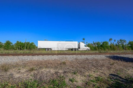 "Boundless routes A colossal 18-wheeler tractor-trailer traverses the highway, as railroad tracks stretch in the foreground against the backdrop of a brilliant blue sky, embodying the endless possibilities of travel and exploration. 