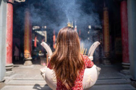 Foto de Vietnamese girl in traditional long dress or Ao Dai dress is praying with incense stick in the burning pot of a pagoda in Ho Chi Minh City, Vietnam. Tet holiday and New Year. . Tet holiday and New Year. Travel concept. - Imagen libre de derechos