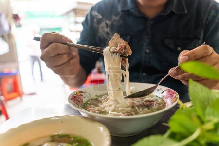 A man using chopsticks and spoon eating traditional Pho Bo vietnamese soup with beef and rice noodles on a metal table, real scene in local restaurant, selective focus