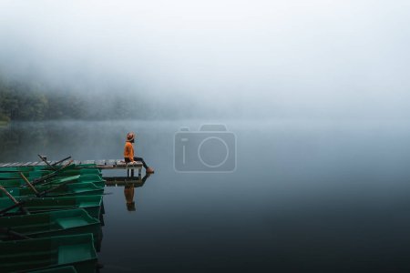 Téléchargez les photos : Side view of fashioned young woman sitting on wooden dock looking at view on a misty morning. Female hipster relaxes on the edge of jetty - en image libre de droit