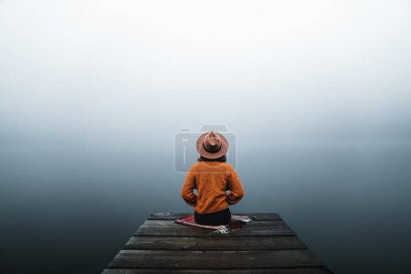 Téléchargez les photos : Back view of fashioned young woman sitting on wooden dock looking at view on a misty morning. Female hipster with brown hat relaxes on the edge of jetty admiring foggy lake. Wonderful nature getaway - en image libre de droit