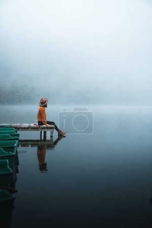 Téléchargez les photos : Side view of fashioned young woman sitting on wooden dock looking at view on a misty morning. Female hipster relaxes on the edge of jetty admiring foggy landscape. Wonderful nature getaway - en image libre de droit