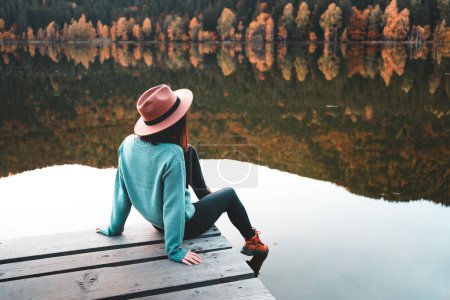 Foto de Side view of fashioned young woman sitting on wooden dock looking at view with beautiful autumn colors. Female hipster with brown hat relaxes on the edge of jetty. Wonderful nature getaway - Imagen libre de derechos