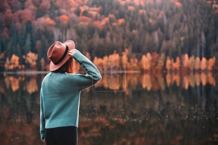 Foto de Side view of young woman with brown hat looking at view against lake, reflections, forest and autumn colors. - Imagen libre de derechos