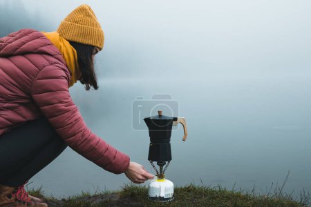Téléchargez les photos : Woman hiker preparing coffee from moka pot coffee maker on camping gas stove by the lake. Adventure, travel, hiking and camping concept - en image libre de droit