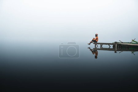 Téléchargez les photos : Side view of fashionable young woman standing on wooden dock looking at view on a misty morning. Female hipster relaxes on the edge of jetty admiring foggy landscape. Wonderful nature getaway - en image libre de droit