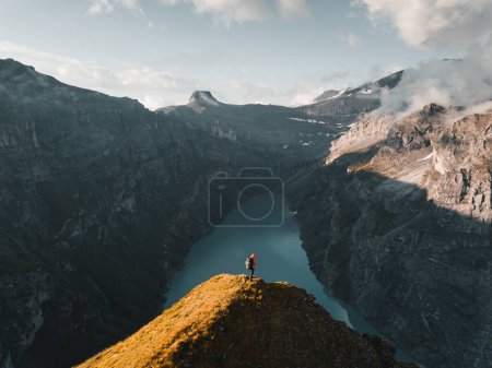 Téléchargez les photos : High Angle back view of hiker in red jacket standing on top of mountain and looking at Limmernsee Lake. Man sitting on mountain edge and enjoying the beautiful view in Glarus, Switzerland - en image libre de droit