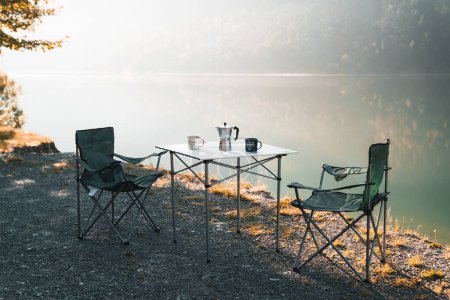 Foto de Al aire libre dos sillas vacías con mesa de picnic y café moka pot para acampar contra el lago tranquilo por la mañana. Equipo para acampar cocina, cocinar en la naturaleza, café al aire libre con ustensils camping - Imagen libre de derechos
