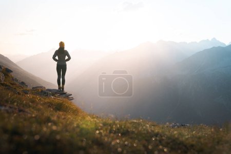 Photo for Young woman hiker standing on the edge of cliff at sunrise. Female tourist reaching summit enjoying amazing sunrise in the mountains.Backlight sunlight with beautiful lens flare and sunbeam.Copy Space - Royalty Free Image