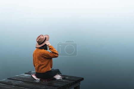 Téléchargez les photos : Side view of fashioned young woman sitting on wooden dock looking at view on a misty morning. Female hipster with brown hat relaxes on the edge of jetty admiring foggy lake. Wonderful nature getaway - en image libre de droit