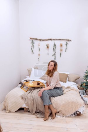 Modern middle aged woman sitting on bed in scandinavian style Christmas. Girl is smiling, wearing neutral color sweater, gray pants. On a white wall, wooden snag decorated with garland, Christmas tree
