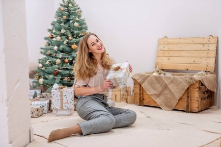 Stylish, modern millennial woman sits at the Artificial Christmas tree, holds a gift in her hands, smiles. Red lipstick on lips. Festive eco-friendly interior. Copy space, close up potrtait