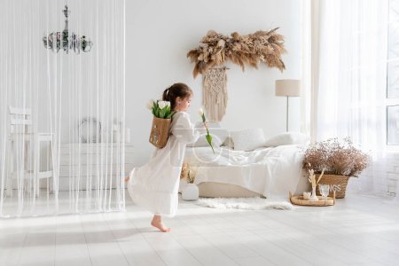 Photo for Little girl in white dress, nightgown dancing with back, faceless, on shoulders is hand-made backpack made of raffia with tulips. Light airy, spring interior in rustic style. Pampas grass on the wall - Royalty Free Image