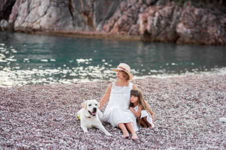 Photo for Pregnant mother with little daughter are sitting along the sand of the beach near the sea with golden retriever. Millennial woman holds dog on leash, girl by the hand. Concept of traveling with pets - Royalty Free Image